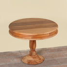 A round wooden pedestal table with a multi-toned natural wood top and turned base, set against a plain beige wall and stone floor.
