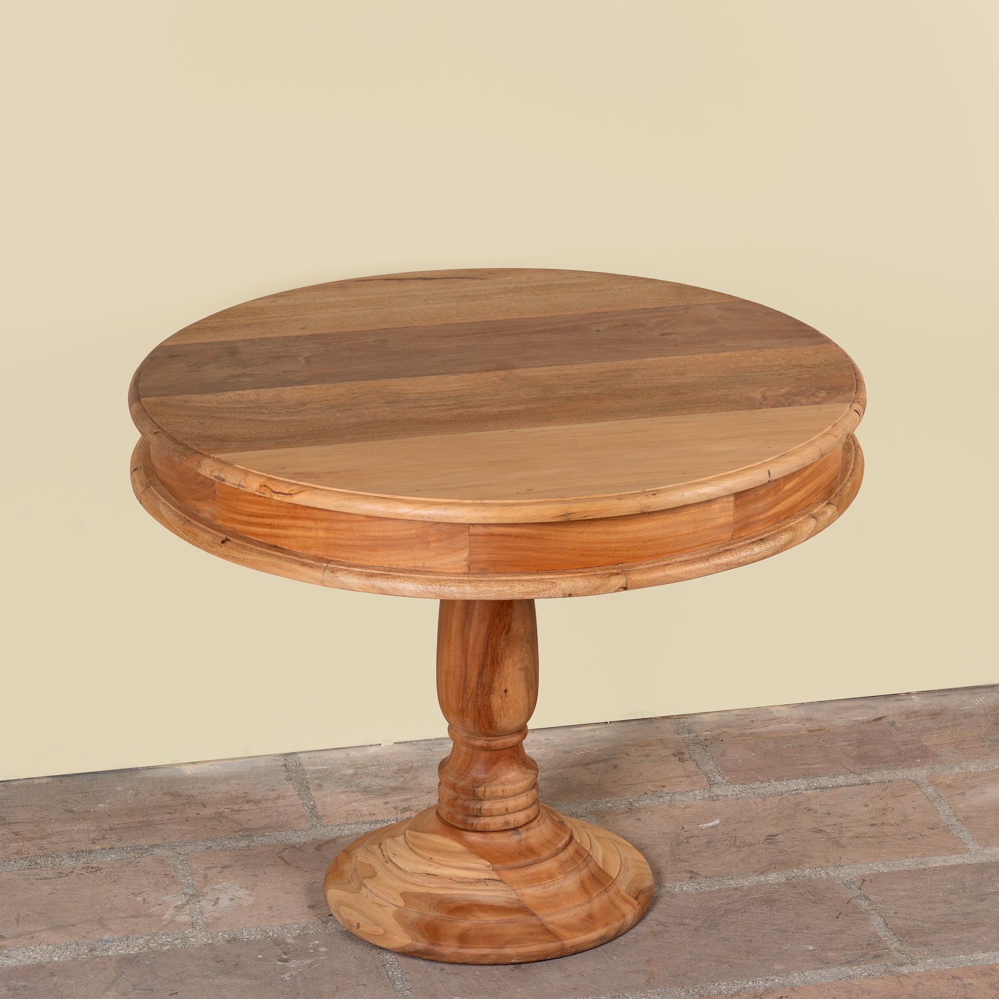 A round wooden pedestal table with a multi-toned natural wood top and turned base, set against a plain beige wall and stone floor.
