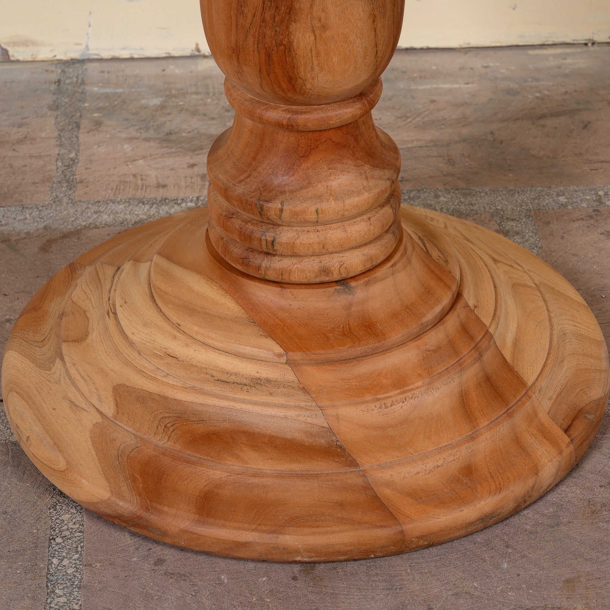 Close-up of a round wooden table base with a turned pedestal column, made from natural wood with visible grain and a warm honey-brown finish, set on a stone floor.