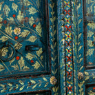 Close up image of a vintage wall panel crafted from an old teak wood window frame, hand-painted with detailed floral designs in cream, red, and green on a rich teal background, featuring arched upper panels and decorative metal ring pulls.
