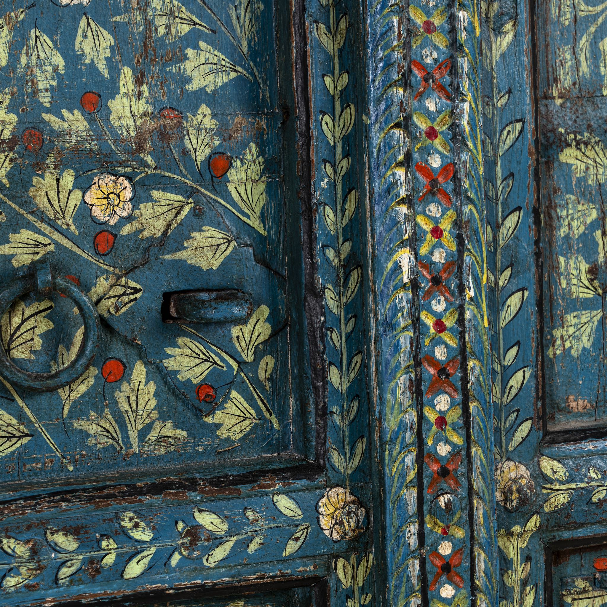 Close up image of a vintage wall panel crafted from an old teak wood window frame, hand-painted with detailed floral designs in cream, red, and green on a rich teal background, featuring arched upper panels and decorative metal ring pulls.

