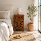 Lifestyle scene with a mango wood night stands with hand carved floral and geometric detailing, warm teak finish; elegant hand carved furniture, 45 × 35 × 65 cm.