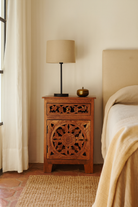 Lifestyle scene with a pair of mango wood night stands with hand carved floral and geometric detailing, warm teak finish; elegant hand carved furniture, 45 × 35 × 65 cm.