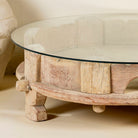 Close up image of a round reclaimed teak coffee table with glass top and hand-carved base; rustic vintage design, 84 cm diameter, 28 cm height.