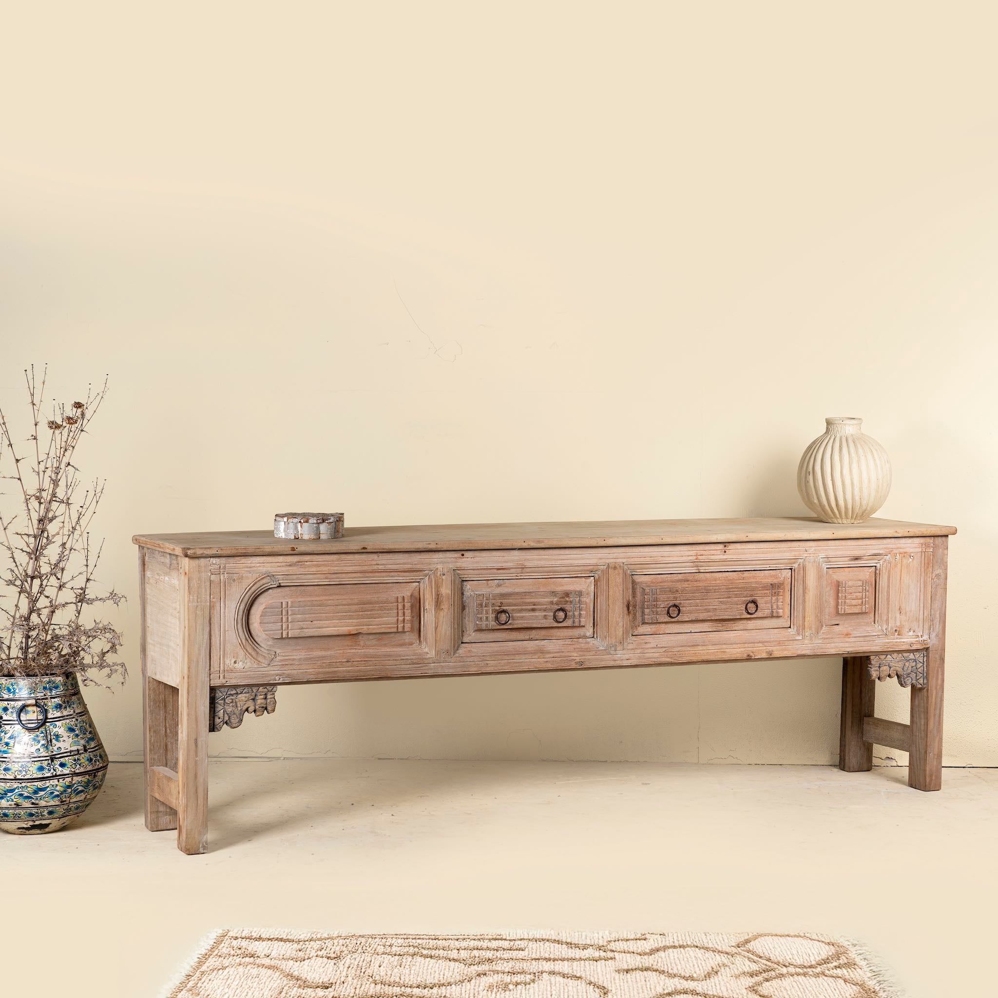 Side angle picture of a antique console table crafted from aged teak wood, featuring detailed hand carvings, two central drawers, and a weathered patina for timeless character.