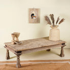 Rustic coffee table Amana made from old reclaimed teak wood, featuring a weathered finish, turned legs, decorative carved apron, and visible metal rivets, styled with earthy decor in a minimal interior setting.
