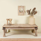 Life style image of the rustic coffee table Amana made from old reclaimed teak wood, featuring a weathered finish, turned legs, decorative carved apron, and visible metal rivets, styled with earthy decor in a minimal interior setting.
