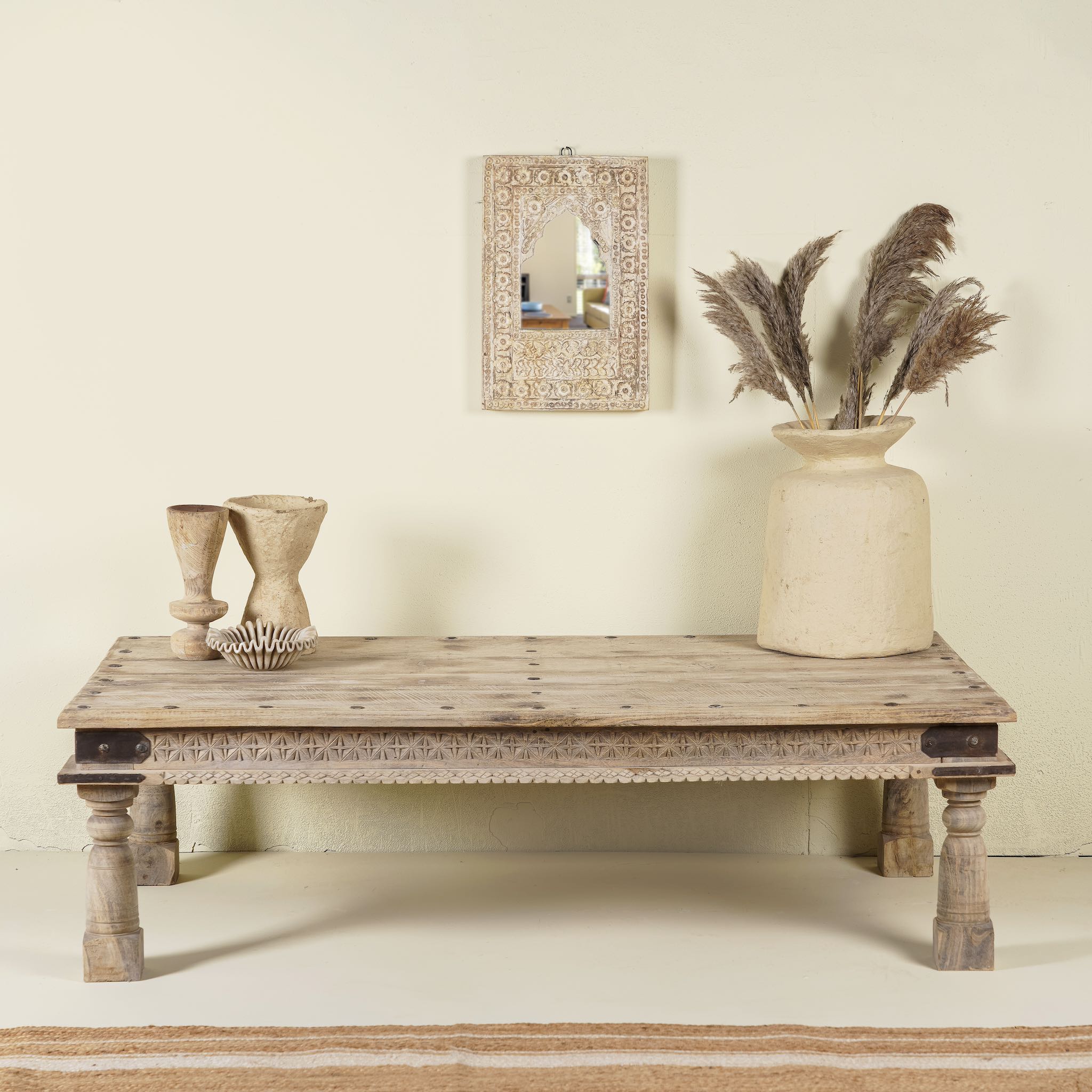 Life style image of the rustic coffee table Amana made from old reclaimed teak wood, featuring a weathered finish, turned legs, decorative carved apron, and visible metal rivets, styled with earthy decor in a minimal interior setting.
