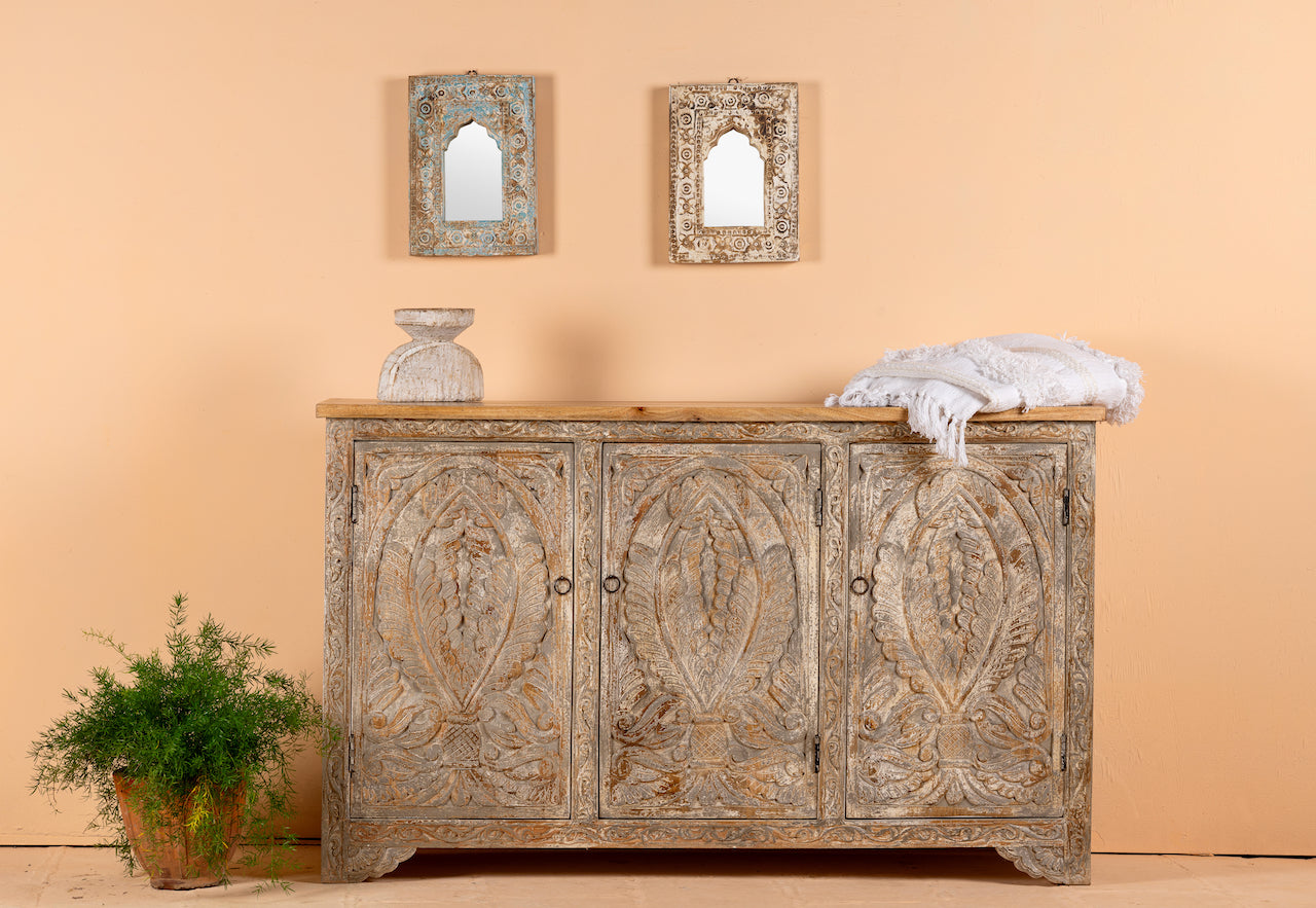 A hand-carved mango wood vintage sideboard with three doors, featuring intricate floral carvings, placed against a neutral backdrop with decorative mirrors above it. A plant and a decorative object are also visible to the side.