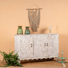 Side angle image of a hand-carved mango wood sideboard with peacock and foliage motifs, whitewashed finish; perfect for Indian home decor, 150 × 95 × 40 cm.