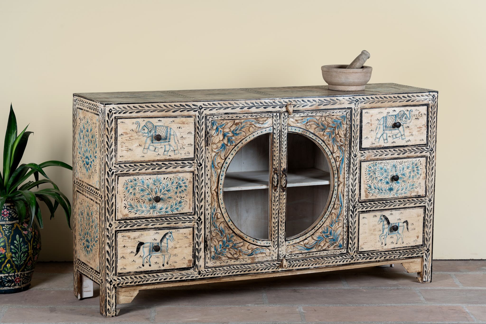 Side angle image of a hand-painted sideboard Anaya made from reclaimed wood with six drawers and two glass doors, featuring detailed animal and floral motifs. A sustainable piece of boho-style artisanal furniture with colourful heritage design.