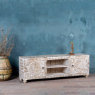 Side angle image of a white washed mango wood lowboard with carved floral doors and open shelving, 180 × 40 × 60 cm.