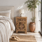 Lifestyle scene with a mango wood bedside table with floral and geometric carvings; natural finish mango wood furniture ideal for bedroom furniture, 45 × 65 × 35 cm.
