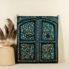 Vintage wall panel crafted from an old teak wood window frame, hand-painted with detailed floral designs in cream, red, and green on a rich teal background, featuring arched upper panels and decorative metal ring pulls.
