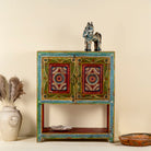 Front view image of a small hand-painted console made from reclaimed wood, featuring vibrant decorative motifs in red, green, blue, and gold on double doors, with intricate floral and geometric patterns and a lower open shelf for added display.
