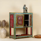 Side angle image with open door of a small hand-painted console made from reclaimed wood, featuring vibrant decorative motifs in red, green, blue, and gold on double doors, with intricate floral and geometric patterns and a lower open shelf for added display.
