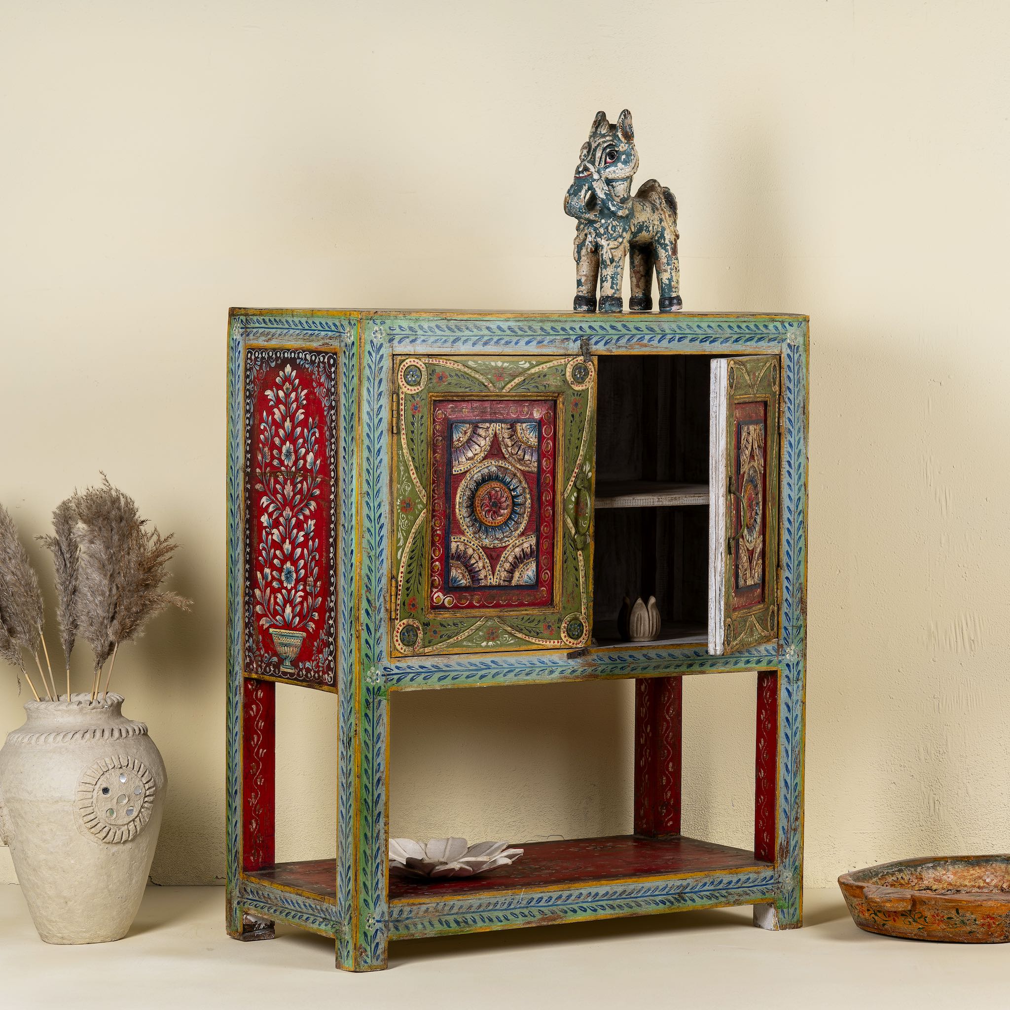Side angle image with open door of a small hand-painted console made from reclaimed wood, featuring vibrant decorative motifs in red, green, blue, and gold on double doors, with intricate floral and geometric patterns and a lower open shelf for added display.

