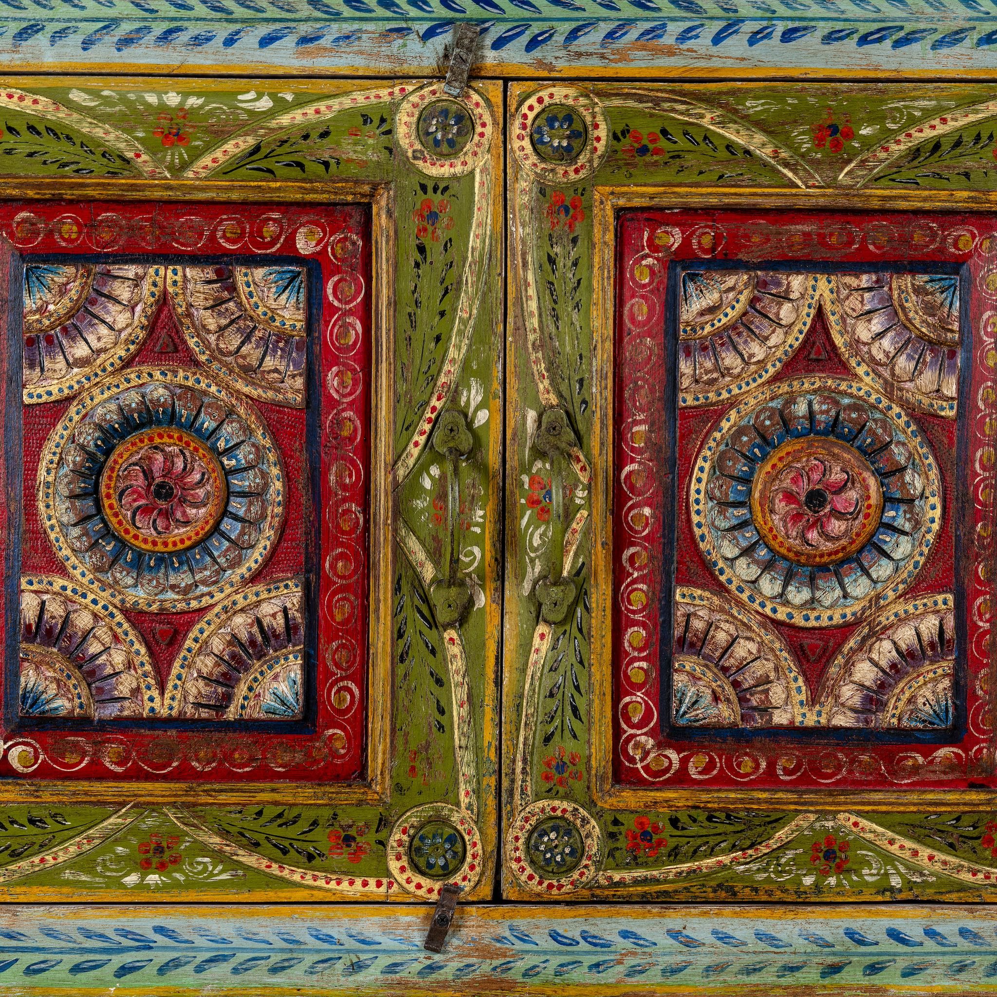 Close up image of a small hand-painted console made from reclaimed wood, featuring vibrant decorative motifs in red, green, blue, and gold on double doors, with intricate floral and geometric patterns and a lower open shelf for added display.
