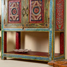 Close up image of a leg of a small hand-painted console made from reclaimed wood, featuring vibrant decorative motifs in red, green, blue, and gold on double doors, with intricate floral and geometric patterns and a lower open shelf for added display.
