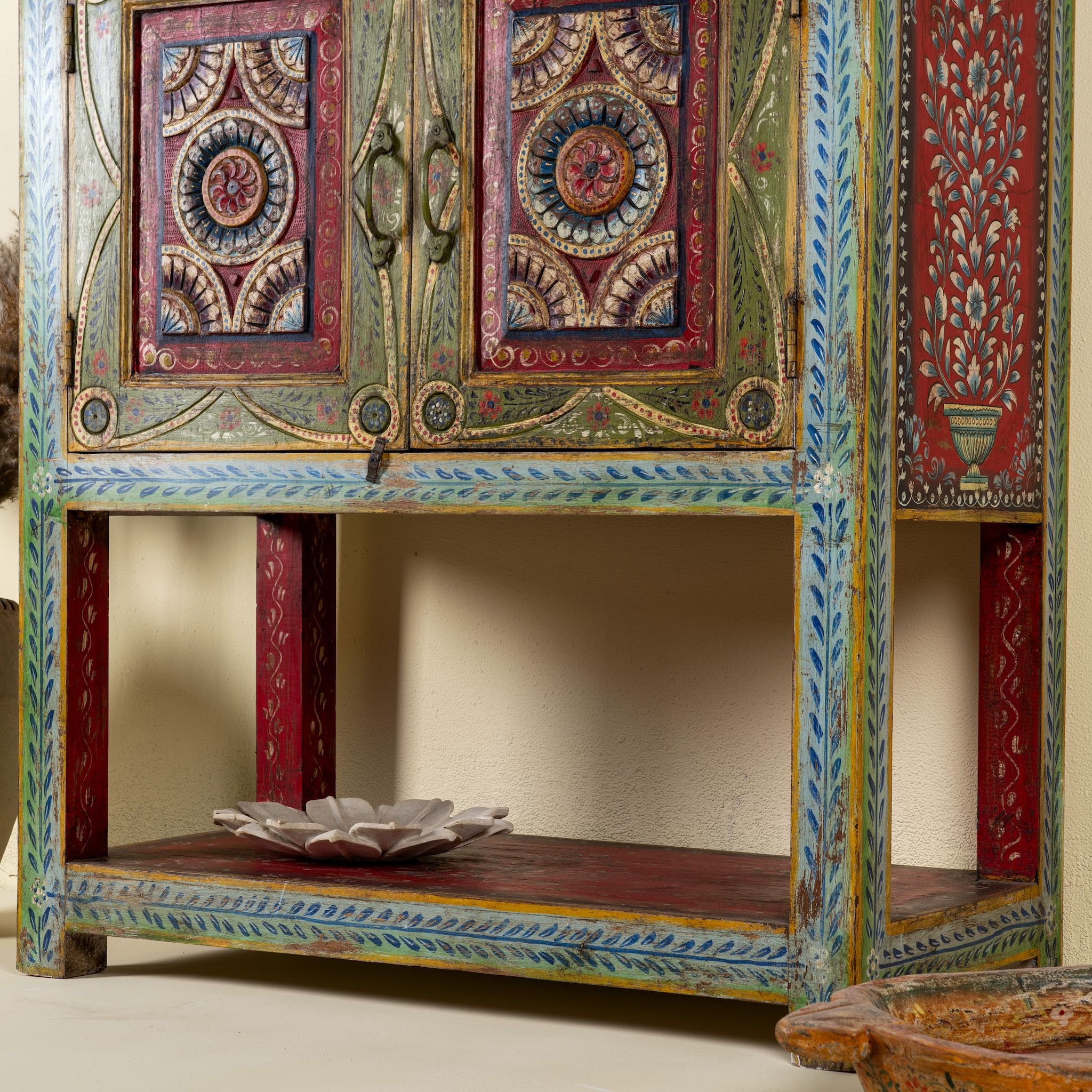 Close up image of a leg of a small hand-painted console made from reclaimed wood, featuring vibrant decorative motifs in red, green, blue, and gold on double doors, with intricate floral and geometric patterns and a lower open shelf for added display.
