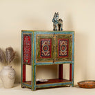 Side angle image of a small hand-painted console made from reclaimed wood, featuring vibrant decorative motifs in red, green, blue, and gold on double doors, with intricate floral and geometric patterns and a lower open shelf for added display.
