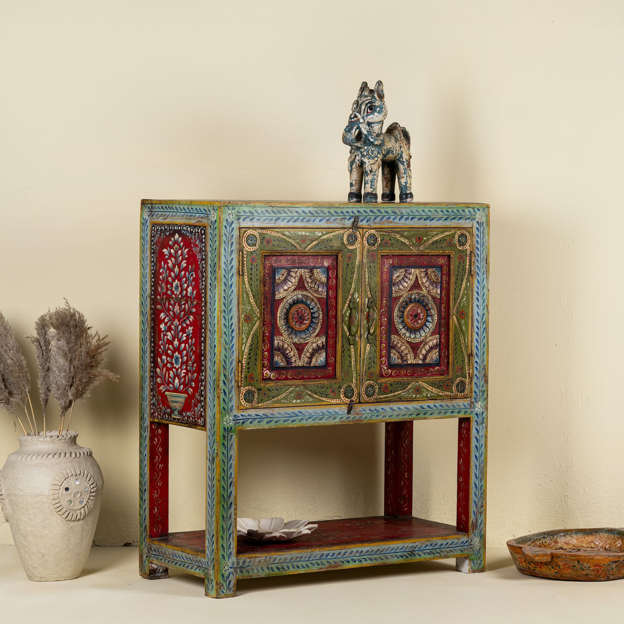 Side angle image of a small hand-painted console made from reclaimed wood, featuring vibrant decorative motifs in red, green, blue, and gold on double doors, with intricate floral and geometric patterns and a lower open shelf for added display.
