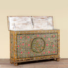 A distressed vintage wooden chest with a hinged lid, featuring hand-painted floral patterns in pink, red, and green on a weathered wood base, set against a plain beige wall.