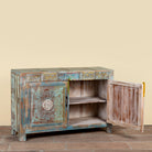 A rustic hand-painted wooden side cabinet with a distressed teal, green, and brown finish, featuring carved floral details on the door and drawers, with one door open to reveal shelves inside.