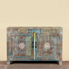 A rustic, hand-painted wooden sideboard cabinet with distressed teal, brown, and gold tones. The cabinet features two doors with ornate circular medallion carvings, decorative panels above, and a distressed finish.