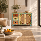 A vibrant, hand-painted wooden sideboard cabinet in a styled living room. The cabinet features colorful floral patterns, woven cane panels on the doors, and turquoise painted legs, set against a neutral beige background with a coffee table and plants.