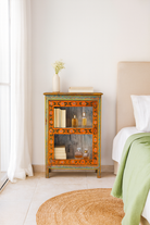 Lifestyle imagination of a vintage display cabinet in recycled wood with hand-painted decorative panels and glass display doors, 57 × 41 × 80 cm.