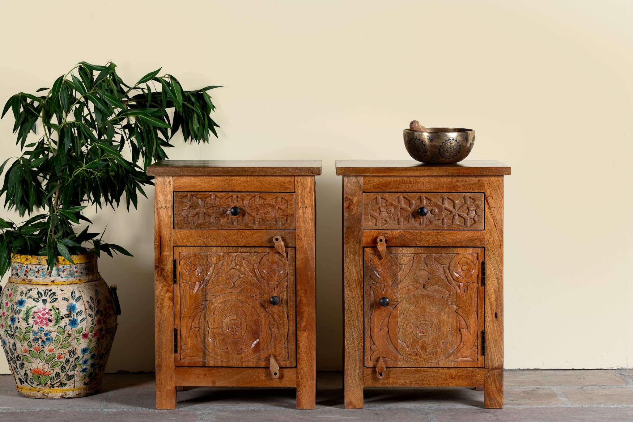 Lifestyle image of a pair of handcrafted bedside tables Avi made from solid mango wood, featuring floral hand-carved motifs inspired by Ibiza style. Each table includes a drawer and a cabinet, with a natural wood finish that highlights artisanal detail and organic warmth.