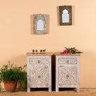 A pair of hand-carved nightstands made from mango wood with a light grey-washed finish, featuring floral patterns. They have multiple storage compartments and are placed in a room with decorative items on and around them.