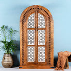 A hand-carved mango wood wardrobe with half-round shape and whitewashed lattice-style panels, featuring intricate carvings. The wardrobe is positioned against a light blue wall, with a potted plant and a brown textured object to the side.