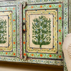 Close up side angle image of a hand-painted sideboard made from reclaimed wood, featuring three drawers and three cabinet doors adorned with intricate floral borders and tree motifs in earthy green and warm tones, finished in a soft distressed patina for a vintage artisanal look.
