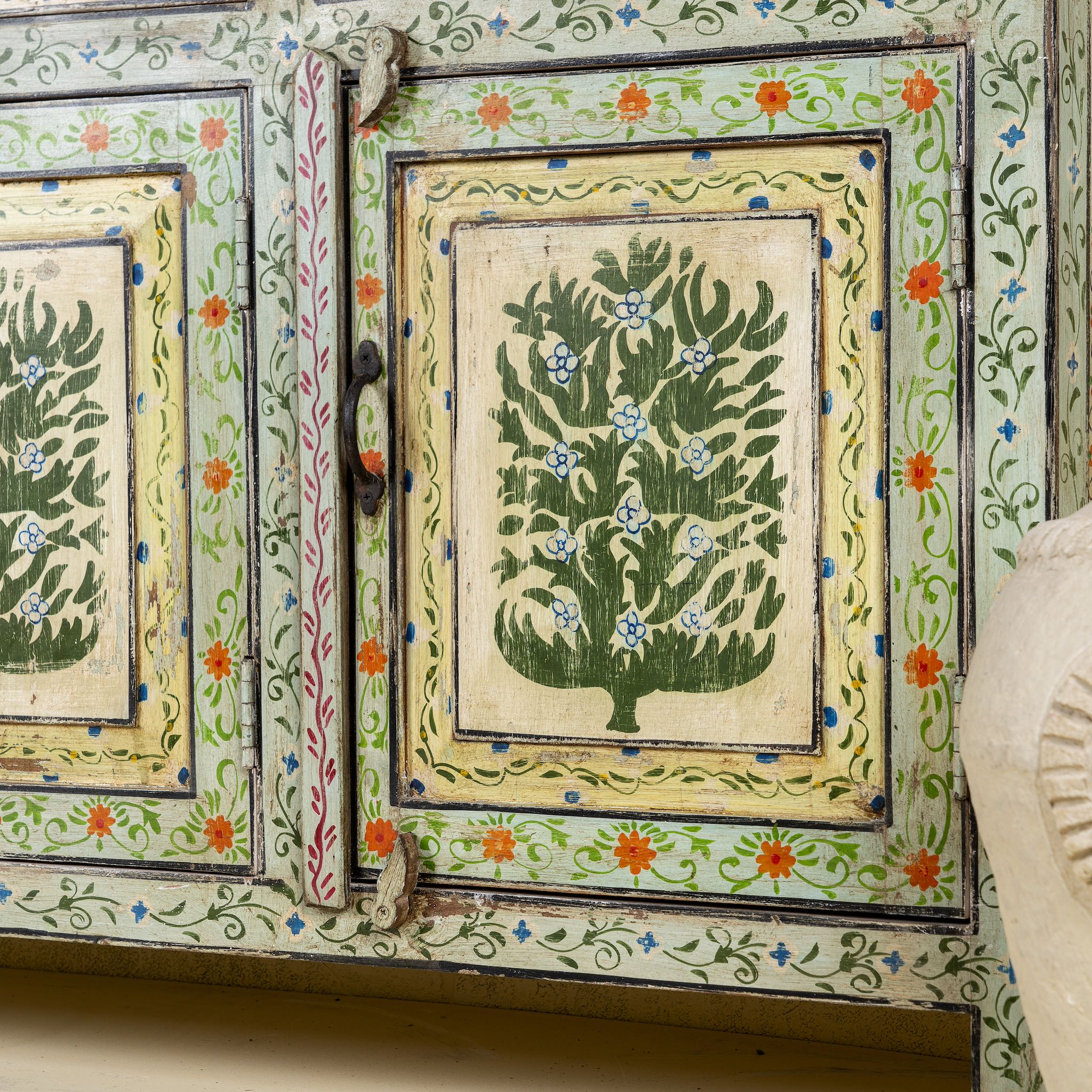 Close up side angle image of a hand-painted sideboard made from reclaimed wood, featuring three drawers and three cabinet doors adorned with intricate floral borders and tree motifs in earthy green and warm tones, finished in a soft distressed patina for a vintage artisanal look.
