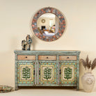Front view image of a hand-painted sideboard made from reclaimed wood, featuring three drawers and three cabinet doors adorned with intricate floral borders and tree motifs in earthy green and warm tones, finished in a soft distressed patina for a vintage artisanal look.
