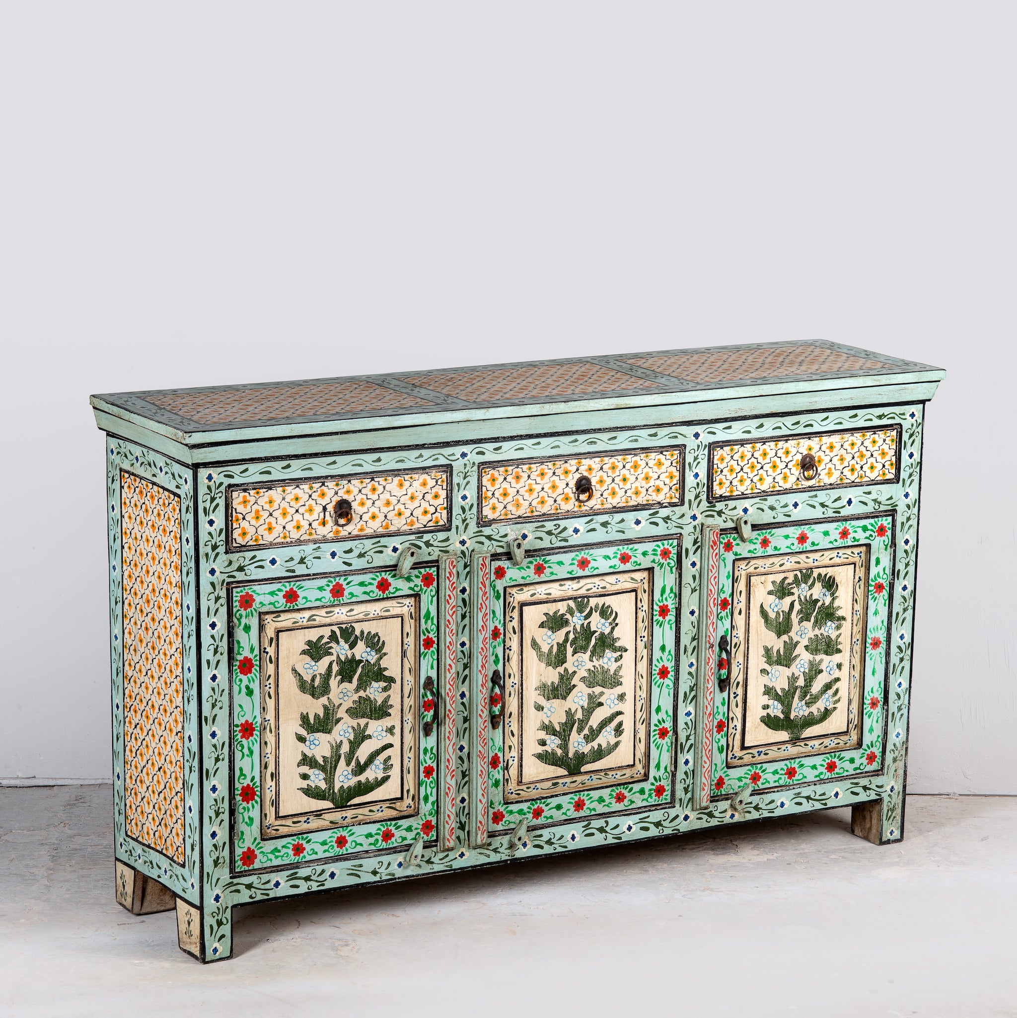 Side angle image of a hand-painted sideboard made from reclaimed wood, featuring three drawers and three cabinet doors adorned with intricate floral borders and tree motifs in earthy green and warm tones, finished in a soft distressed patina for a vintage artisanal look.
