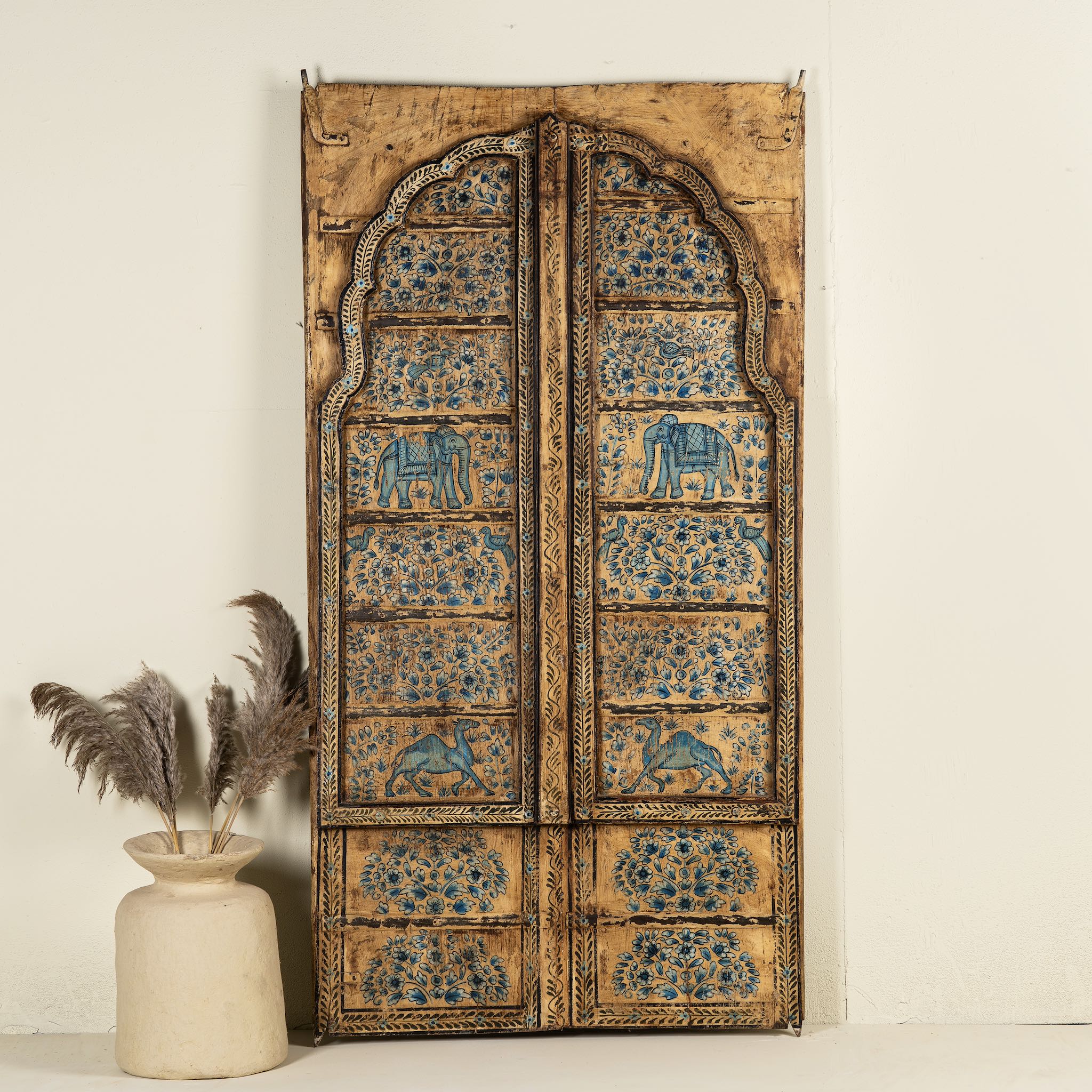 Hand-painted wooden wall panel made from an old window frame, featuring intricate blue floral patterns along with stylised animal motifs such as elephants and camels, set against a distressed golden-brown background with an arched top frame.
