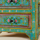 Close up image of the leg part of a  hand-painted reclaimed wood nightstand with vibrant colours and floral patterns, featuring three drawers, placed next to a vase with dried plants.