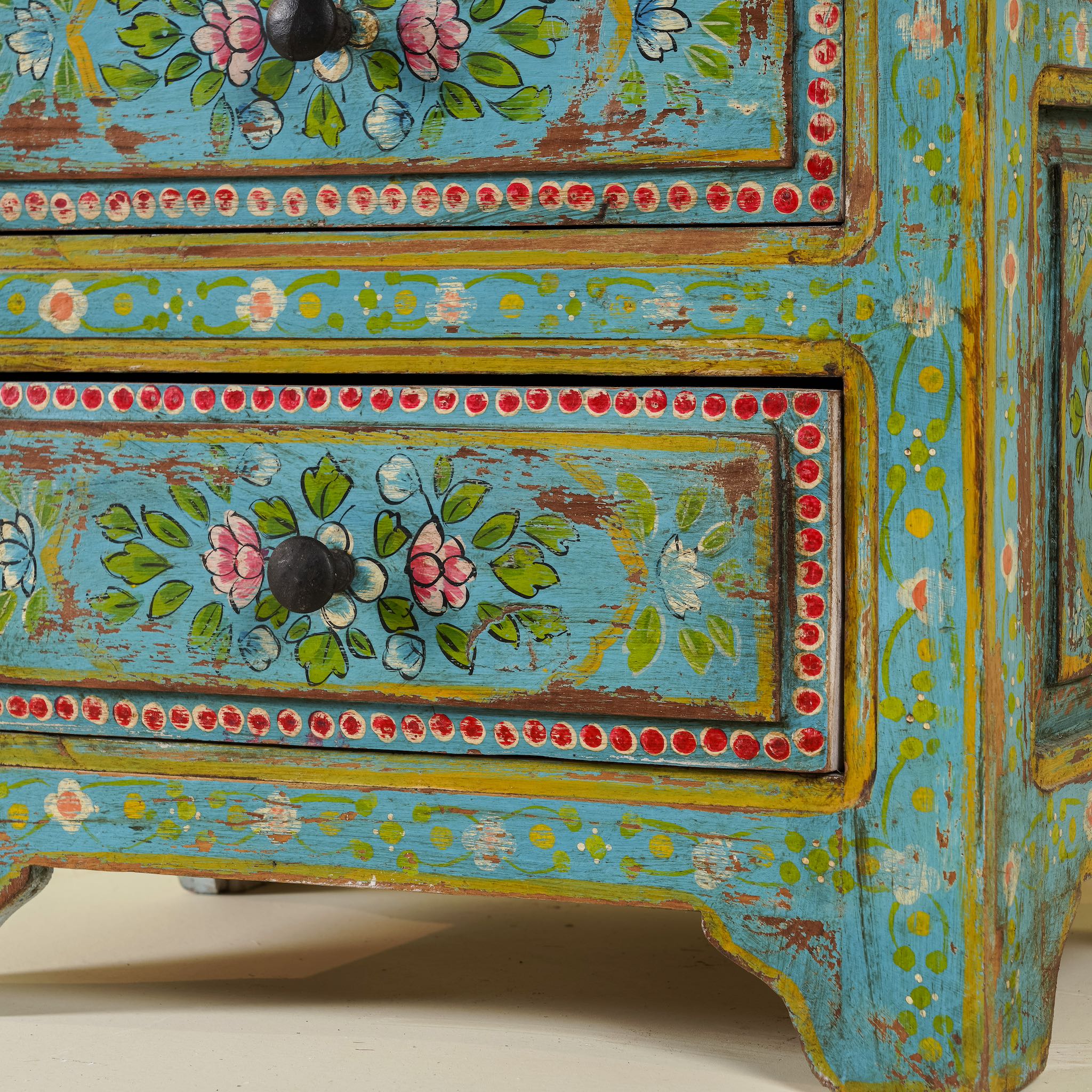 Close up image of the leg part of a  hand-painted reclaimed wood nightstand with vibrant colours and floral patterns, featuring three drawers, placed next to a vase with dried plants.
