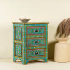Side angle image of a  hand-painted reclaimed wood nightstand with vibrant colours and floral patterns, featuring three drawers, placed next to a vase with dried plants.