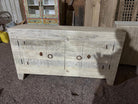 A carved wooden sideboard cabinet in a weathered off-white finish, featuring circular and floral engravings and rustic metal ring pulls, photographed in a workshop or warehouse setting.