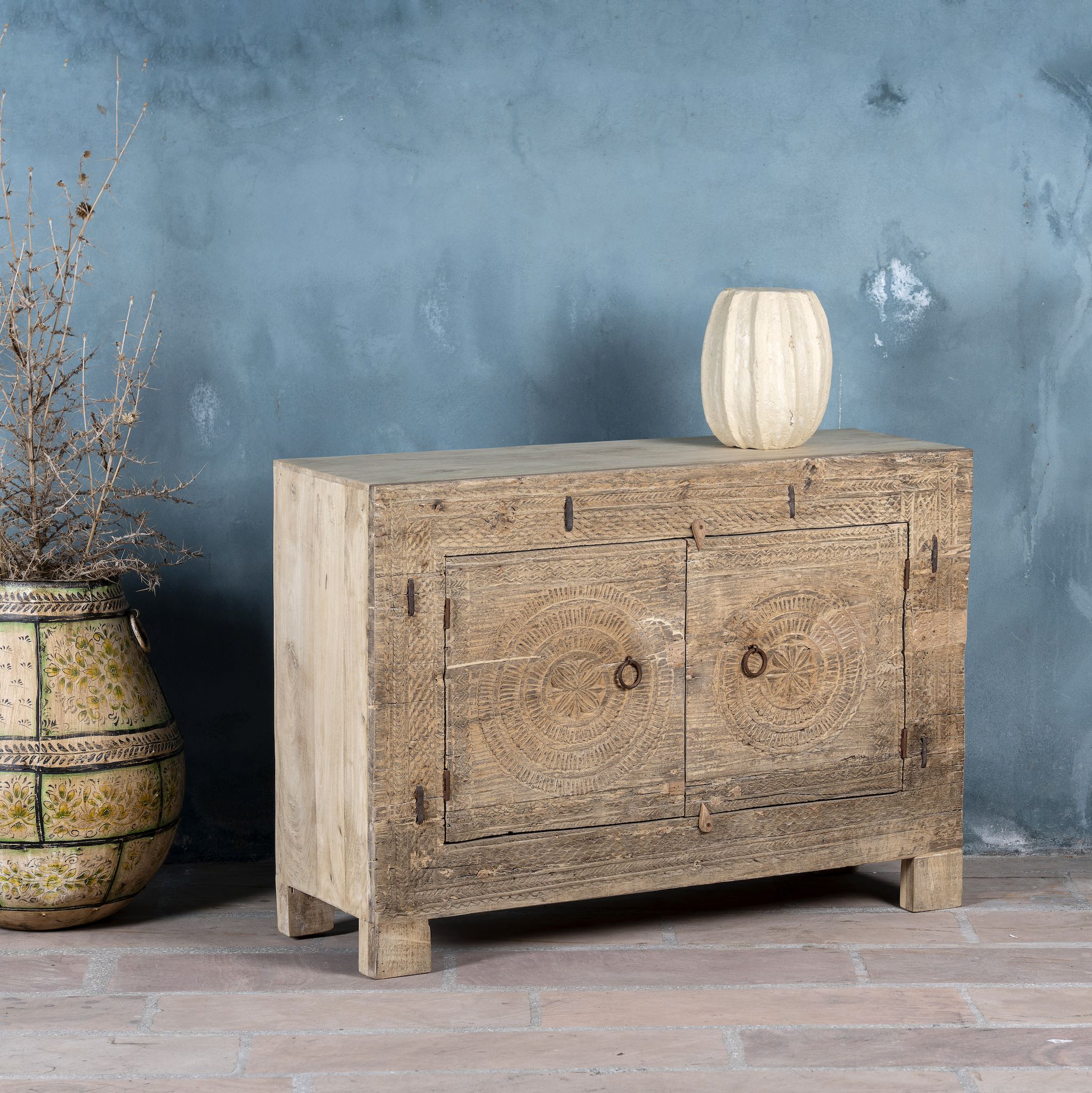 Side angle of a sideboard with hand-carved teak wood front, sandblasted for a sun-worn finish- a unique piece in rustic Ibiza style with circular mandala carvings and iron ring handles.