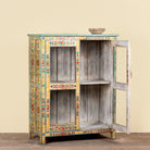 A rustic hand-painted wooden cabinet with a distressed gray wood finish, decorated with colorful floral patterns in yellow, green, red, and blue. The cabinet features two doors, one of which is open, revealing two interior shelves, and is set against a plain beige wall.