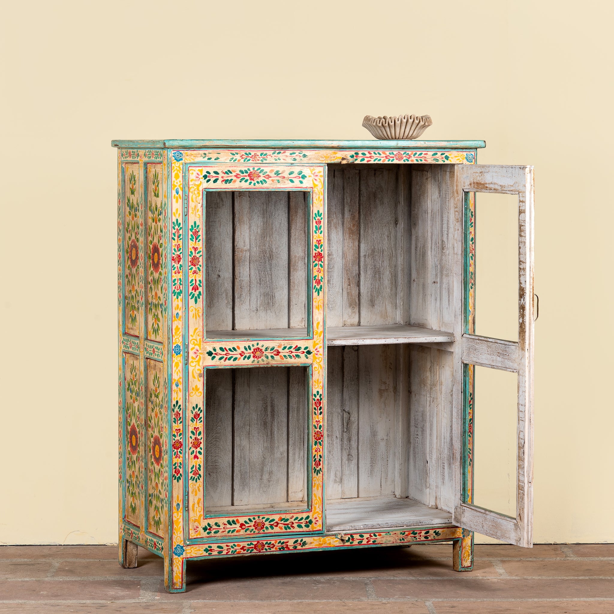 A rustic hand-painted wooden cabinet with a distressed gray wood finish, decorated with colorful floral patterns in yellow, green, red, and blue. The cabinet features two doors, one of which is open, revealing two interior shelves, and is set against a plain beige wall.