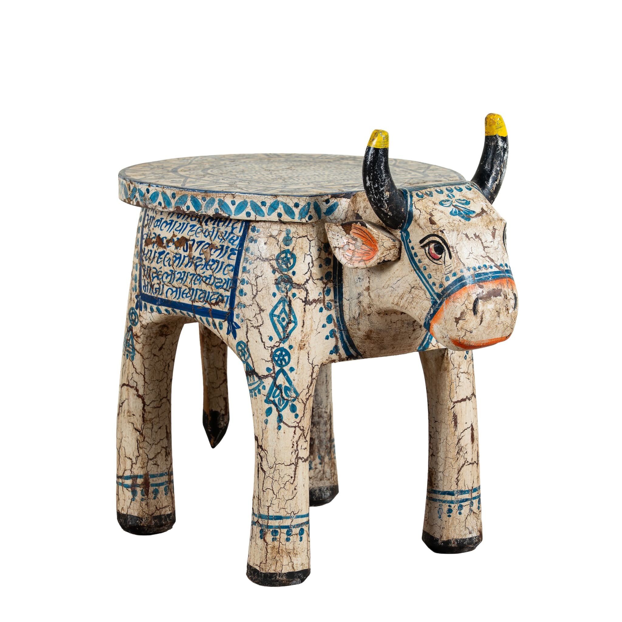 White background image of a hand-painted wooden cow-shaped side table with Indian script and blue folk art patterns, perfect for vintage and India-inspired home interiors.