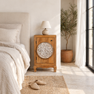 Lifestyle scene with a whitewashed mango wood nightstand with hand-carved floral door – a handcrafted example of carved wooden furniture ideal for rustic and vintage bedrooms.