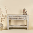 Front view image of a handcarved console table made from reclaimed teak wood, featuring a detailed front panel with geometric floral motifs and a distressed whitewashed finish, complemented by a lower shelf for added storage or display.
