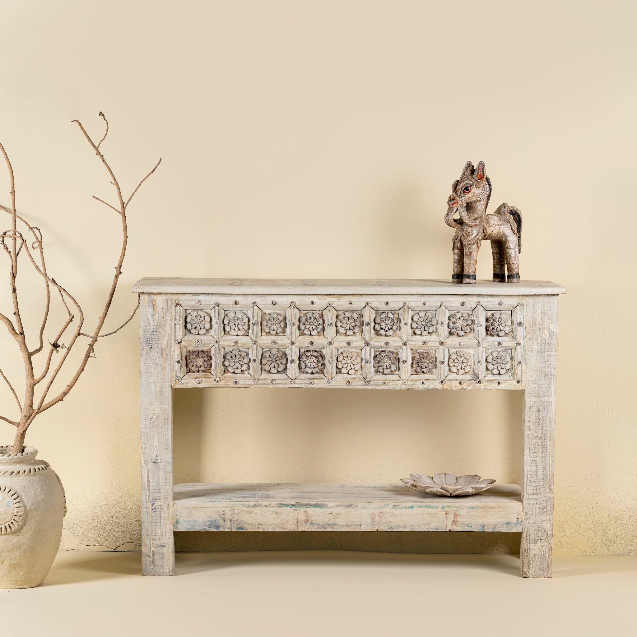 Front view image of a handcarved console table made from reclaimed teak wood, featuring a detailed front panel with geometric floral motifs and a distressed whitewashed finish, complemented by a lower shelf for added storage or display.
